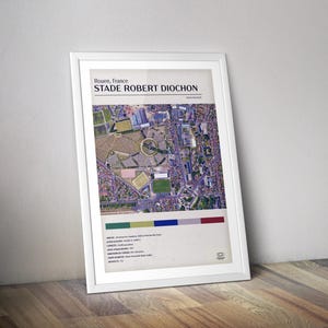 May include: An aerial photograph of Stade Robert Diochon, a soccer stadium in Rouen, France. The stadium is surrounded by a parking lot and residential buildings. The image includes the stadium's name and location.