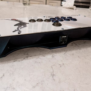 Puede incluir: Un stick de arcade negro con un panel superior blanco que presenta un gráfico de una mujer con un vestido blanco. El stick tiene una base negra con un borde plateado y un panel de botones negro con botones azules. El stick está etiquetado como "Brook" en el lateral.