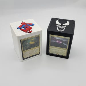 May include: Two card storage boxes. One is white with a red, blue, and black Spider-Man design on the lid. The other is black with a white Venom design on the lid. Each box has a card visible in the front.