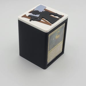 Könnte beinhalten: Schwarz-weiße Kartenaufbewahrungsbox mit weißem Deckel und schwarzem Korpus. Die Oberseite zeigt eine schwarze Silhouette einer Person mit einer Waffe und zwei braunen Holzbrettern. Eine Magic: The Gathering-Karte ist auf der Seite sichtbar.