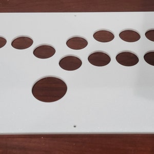 Puede incluir: Un panel de control de arcade blanco con agujeros pre-perforados para botones y un joystick. El panel tiene forma rectangular con un agujero circular grande y varios agujeros circulares más pequeños.