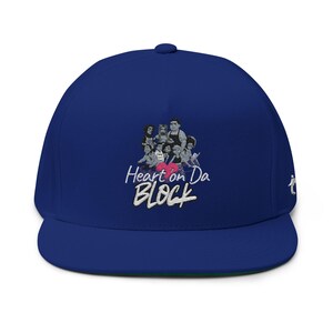 May include: A royal blue baseball cap with a flat brim. The cap features an embroidered design of a group of people and the text "Heart on Da BLOCK" in white and pink.