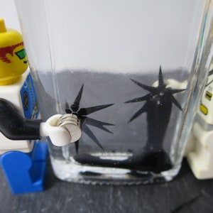 Könnte beinhalten: Eine Lego-Minifigur hält einen Stapel silberner Magnete in der Nähe eines Glasbehälters, der mit einer schwarzen Flüssigkeit gefüllt ist. Die Flüssigkeit enthält mehrere schwarze sternförmige Objekte, die von den Magneten angezogen werden.