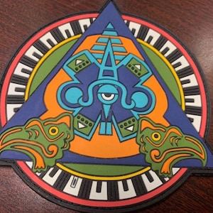 Pode incluir: Um remendo circular colorido com um design azul, laranja e verde. O centro apresenta um olho estilizado com uma forma de pirâmide acima. Duas cabeças de pássaros verdes com bicos laranja estão de cada lado do olho. O círculo externo tem um padrão preto e branco com uma borda vermelha.