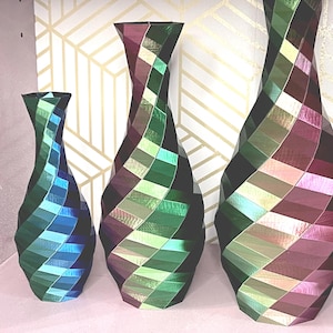 May include: Three decorative vases in varying sizes, featuring a geometric, diamond-patterned design. The vases showcase a color palette of green, blue, and purple hues. The vases are displayed on a pink surface against a gold and white patterned background.