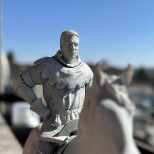May include: A grey 3D printed figurine of a knight riding a horse. The knight is wearing armour and a cloak, and the horse is in a galloping pose.