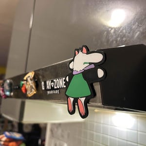 May include: A collection of decorative magnets, including a cartoon character in a green dress and a purple scarf. The text "GAY-ZONE WARFARE" is visible on a metal surface. Other magnets are also visible.