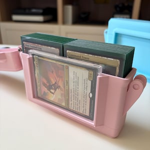 Op de afbeelding: Een lichtroze plastic deckbox met meerdere verzamelkaarten in groene hoesjes. De voorkant van de doos heeft een doorzichtig venster met daarin een kaart met zichtbare tekst, waaronder "Aurelia, the Warleader" en "Legendary Creature - Angel".