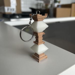 May include: A white and brown 3D printed pagoda keychain. The keychain is made of plastic and has a silver metal ring.