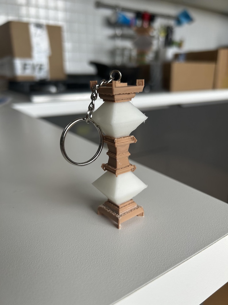 May include: A white and brown 3D printed pagoda keychain. The keychain is made of plastic and has a silver metal ring.