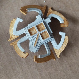 Puede incluir: Un diseño geométrico impreso en 3D, dorado y gris, con un cuadrado en el centro. El diseño recuerda a una placa de circuito.