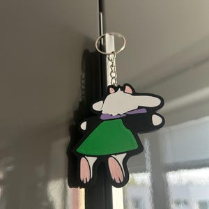 May include: A cartoon-style keychain featuring a character with a white head, purple collar, and green skirt. The keychain is black with pink feet and is attached to a silver keyring.