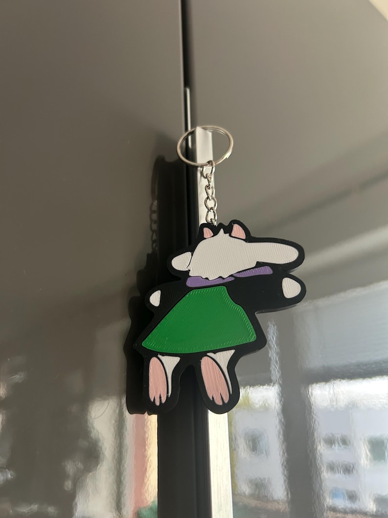 May include: A cartoon-style keychain featuring a character with a white head, purple collar, and green skirt. The keychain is black with pink feet and is attached to a silver keyring.