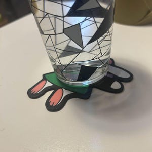 May include: A clear glass with a geometric black, gray, and white design, filled with water, sits on a bunny-shaped coaster. The coaster has a green base with black edges and pink and black bunny feet.