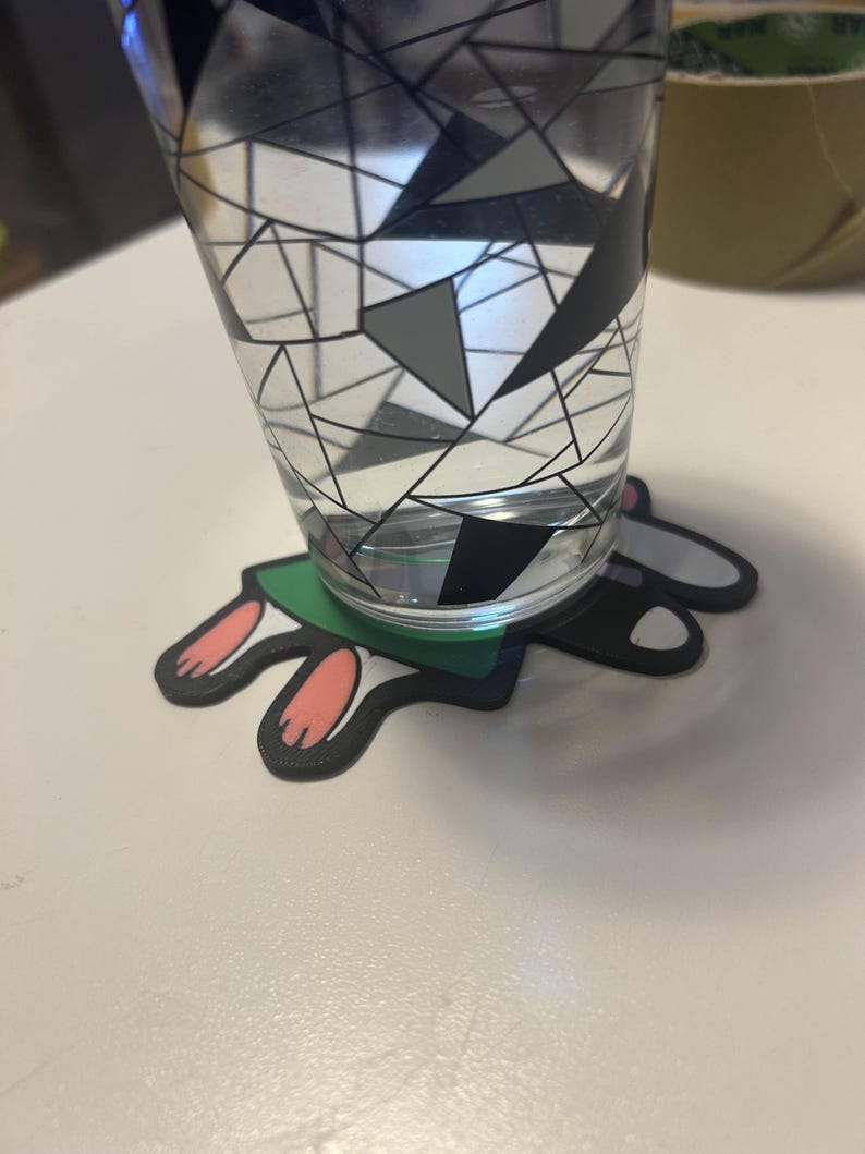 May include: A clear glass with a geometric black, gray, and white design, filled with water, sits on a bunny-shaped coaster. The coaster has a green base with black edges and pink and black bunny feet.