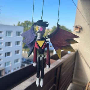 May include: A hanging puppet with a black hat, a yellow triangle, and red and black arms and legs. The puppet has purple and maroon wings with yellow accents. The puppet is suspended by green strings against a blue sky.