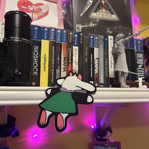 May include: A shelf displays video games, a camera lens, and figurines. Game titles include "Bioshock," "Cyberpunk," and "Dead Cells." A cartoon sticker of a character with a white head, green dress, and pink feet is in the foreground. The shelf is illuminated with purple lights.