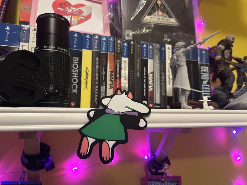 May include: A shelf displays video games, a camera lens, and figurines. Game titles include "Bioshock," "Cyberpunk," and "Dead Cells." A cartoon sticker of a character with a white head, green dress, and pink feet is in the foreground. The shelf is illuminated with purple lights.