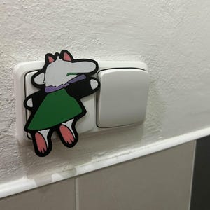 May include: A colorful cartoon character sticker is placed on a white light switch. The sticker features a white creature with a green dress, black arms, and pink feet. The light switch is on a white wall.