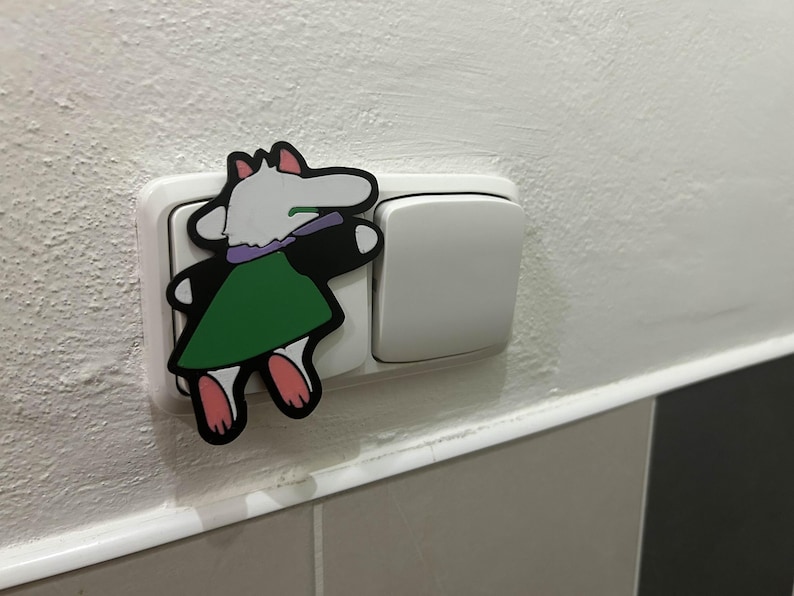 May include: A colorful cartoon character sticker is placed on a white light switch. The sticker features a white creature with a green dress, black arms, and pink feet. The light switch is on a white wall.