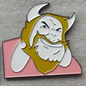 Puede incluir: Un pin de esmalte con una ilustración de dibujos animados de un vikingo con cara y cuernos blancos, barba y pelo amarillos y un fondo rosa. El pin es una representación estilizada de una figura vikinga.