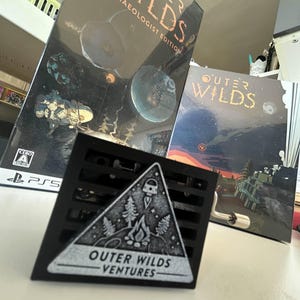 Puede incluir: Un emblema triangular negro y plateado con el texto "OUTER WILDS VENTURES" y un gráfico de una fogata, árboles y un cohete. El emblema está frente a dos cajas de juego con el título "OUTER WILDS" y "ARCHAEOLOGIST EDITION".