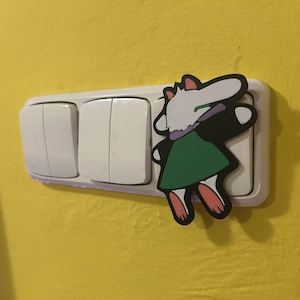 May include: A white light switch with three buttons, with a cartoon character sticker on the right side. The sticker features a white and black character with a green dress and pink feet, on a yellow wall.