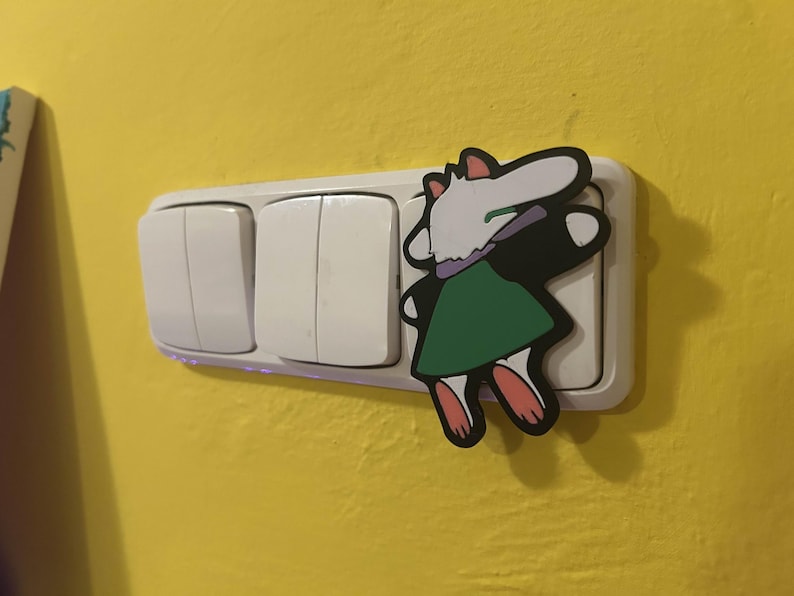 May include: A white light switch with three buttons, with a cartoon character sticker on the right side. The sticker features a white and black character with a green dress and pink feet, on a yellow wall.