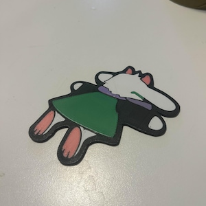 May include: A 3D-printed figurine of a cartoon character, featuring a white face and ears, a green tunic, and black outlines. The character's paws are pink, and the figure is lying down on a white surface.