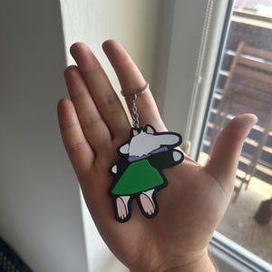 May include: A cartoon-style keychain featuring a character with a white head, a purple collar, and a green dress. The keychain is attached to a silver metal ring. The character's feet are pink, and the outline is black.