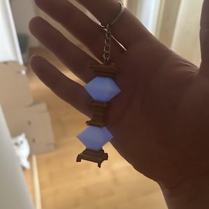 May include: A keychain featuring a three-tiered, light blue, diamond-shaped lantern with a brown wooden base. The lantern is attached to a silver chain and ring.