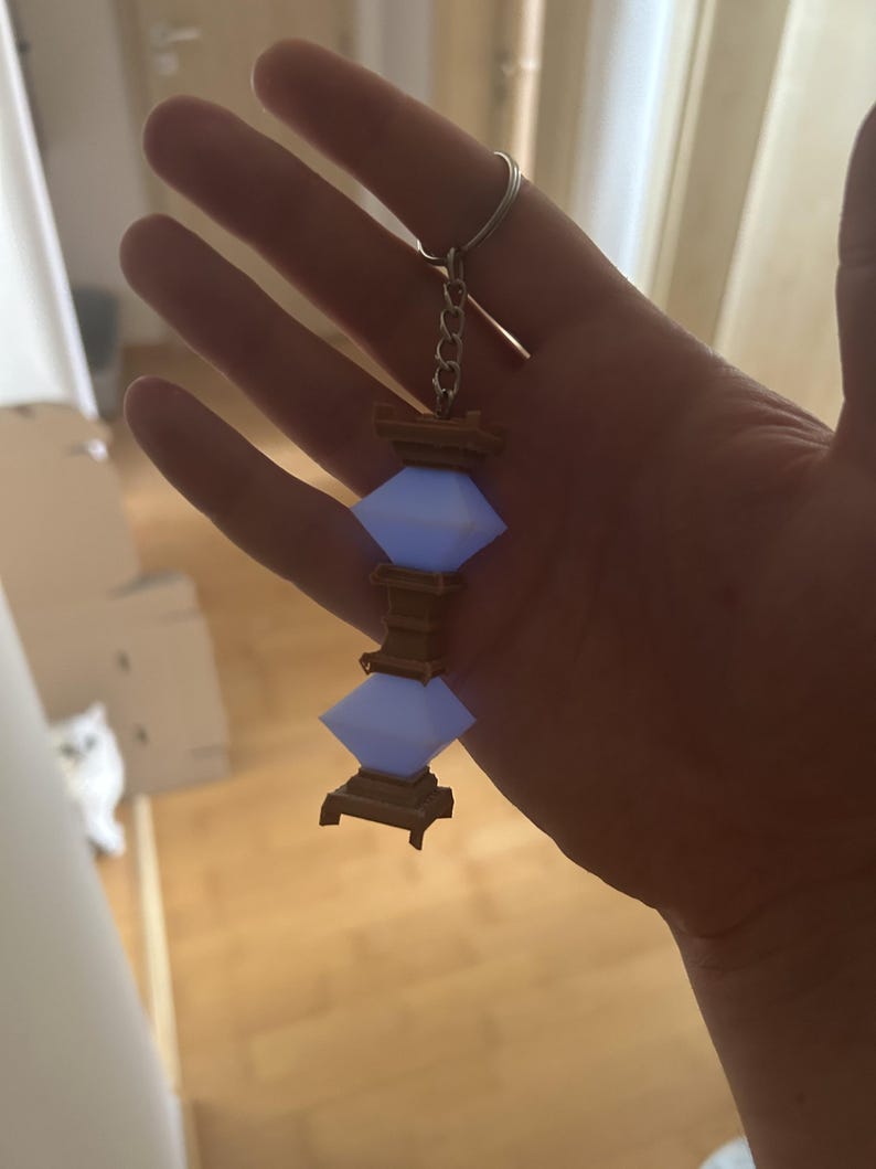 May include: A keychain featuring a three-tiered, light blue, diamond-shaped lantern with a brown wooden base. The lantern is attached to a silver chain and ring.