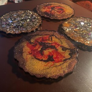 May include: Four resin coasters with glitter and a wood-grain design. The coasters have a dark brown edge and a mix of gold, silver, and red glitter. The center of each coaster has a unique pattern with red and yellow hues.