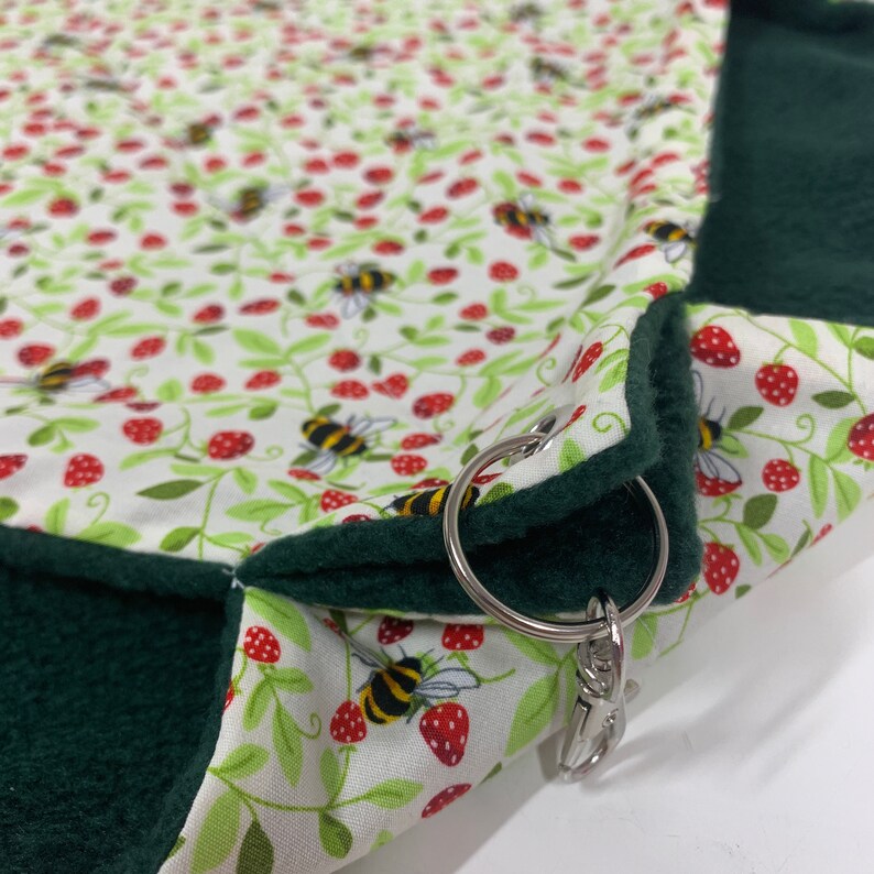 Op de afbeelding: Een groen en wit stoffen dierenbed met een aardbei- en bijenpatroon. Het bed heeft een metalen ring en een clip om op te hangen.