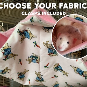 Op de afbeelding: Een roze en wit hangbed van stof voor kleine huisdieren, met een Peter Rabbit-print. Het bed is gemaakt van zacht stof en heeft een roze fleece voering. Een rat rust in het bed. De tekst "CHOOSE YOUR FABRIC! CLASPS INCLUDED" is zichtbaar bovenaan de afbeelding.