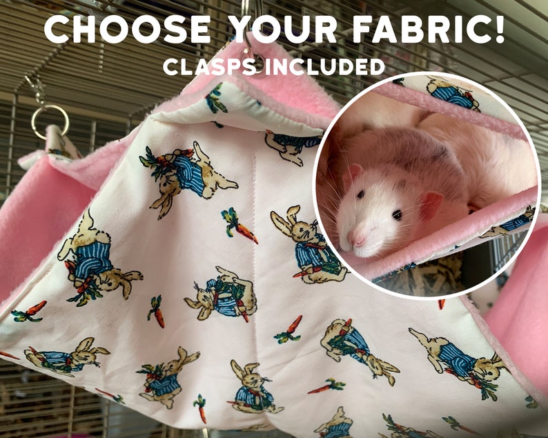 Op de afbeelding: Een roze en wit hangbed van stof voor kleine huisdieren, met een Peter Rabbit-print. Het bed is gemaakt van zacht stof en heeft een roze fleece voering. Een rat rust in het bed. De tekst "CHOOSE YOUR FABRIC! CLASPS INCLUDED" is zichtbaar bovenaan de afbeelding.