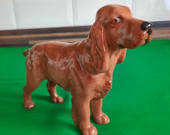 May include: A brown ceramic figurine of a cocker spaniel dog. The dog is standing with its head turned to the right. The figurine is glazed with a brown and white finish.