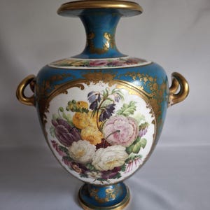 May include: A ceramic vase with a teal body and gold detailing. The vase showcases a central panel with a floral arrangement in pink, yellow, purple, and white. It has two gold handles and a wide, gold-edged top. The vase is a decorative item.