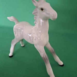 Puede incluir: Figura de cerámica blanca de un potro con detalles grises en las patas y la crin. La figura tiene un acabado brillante y está posada de pie con la cabeza ligeramente girada hacia un lado. Las pezuñas son negras.