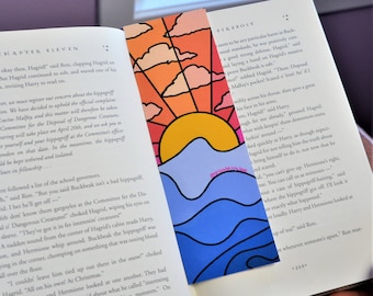 May include: A close-up of a book with a bookmark featuring a sunset over a blue ocean. The bookmark is made of paper and has a colorful design. The book is open to a page with text about a hippogriff.