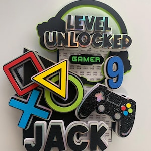 May include: A colorful cake topper with the words "LEVEL UNLOCKED" and "GAMER". It features a controller, a number 9, and the name "JACK". The design includes a PlayStation controller symbol and a triangle, circle, and cross.