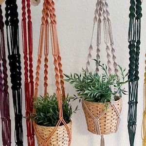 Könnte beinhalten: Sechs Makramee-Pflanzenhänger in verschiedenen Farben, darunter Schwarz, Rot, Orange, Beige, Grün und Gelb. Die Hänger sind aus Baumwollschnur gefertigt und haben ein schlichtes Design. Zwei der Hänger haben Pflanzen.