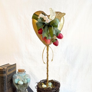 May include: A gold heart-shaped paper mache ornament with a green bow, a butterfly, and red strawberries. The ornament is attached to a gold stick and placed in a gold pot with a keyhole design. The pot is surrounded by a nest with brown twigs and moss, and there are blue speckled eggs inside the nest.
