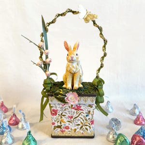May include: A small, white and gold butterfly sits atop a green, twisted wire wreath. A ceramic bunny sits on a floral patterned box with a green ribbon. The box is surrounded by foil-wrapped chocolate kisses.