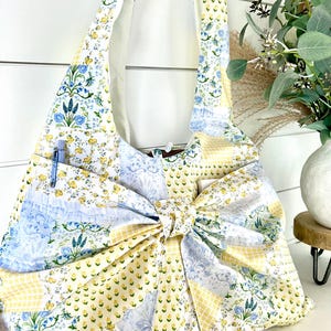 Puede incluir: Bolso de hombro de patchwork con un gran lazo. El bolso presenta estampados florales, de cuadros y lisos en amarillo, azul y blanco. Un bolígrafo azul es visible en el bolsillo lateral. El bolso tiene un estilo suave e informal.