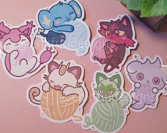Puede incluir: Seis pegatinas de personajes de dibujos animados coloridos con Pokémon sosteniendo ovillos de lana. Los personajes son un Eevee rosa, un Espeon azul, un Flareon rojo, un Sylveon blanco, un Leafeon verde y un Umbreon morado.