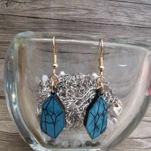 May include: A pair of blue crystal-shaped earrings with gold hooks. The earrings are hanging from a glass container filled with silver chains and beads.