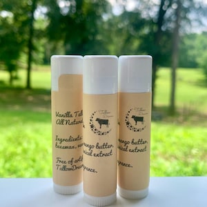 Può includere: Tre tubetti di balsamo per le labbra con tappi bianchi ed etichette beige. Le etichette presentano testo tra cui "Vanilla Tall All Natural" e "mango butter, botanical extract". È visibile anche un'illustrazione di una mucca. Lo sfondo è un paesaggio verde sfocato.