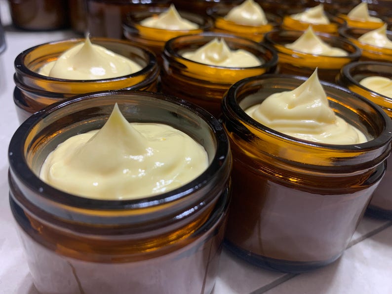 Pode incluir: Close-up de v&aacute;rios pequenos frascos de vidro &acirc;mbar cheios de uma subst&acirc;ncia cremosa amarelo p&aacute;lido. Cada frasco tem um topo arredondado com um redemoinho de creme, criando um pico. Os frascos est&atilde;o dispostos pr&oacute;ximos uns dos outros.