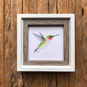May include: A framed watercolor painting of a hummingbird in flight. The hummingbird is green, red, and white with a long beak. The painting is signed "Jenna" in the bottom left corner.